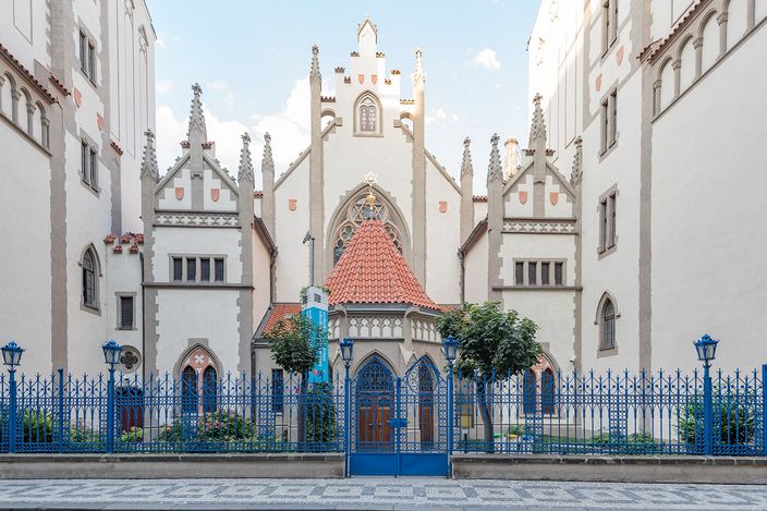 A white neo-Gothic synagogue facade with three stepped gables and slender pinnacles, a small kiosk with a red-tiled pyramid roof standing in front of its central portal, seen behind a blue-painted iron railing with clipped trees on either side in low sunlight.