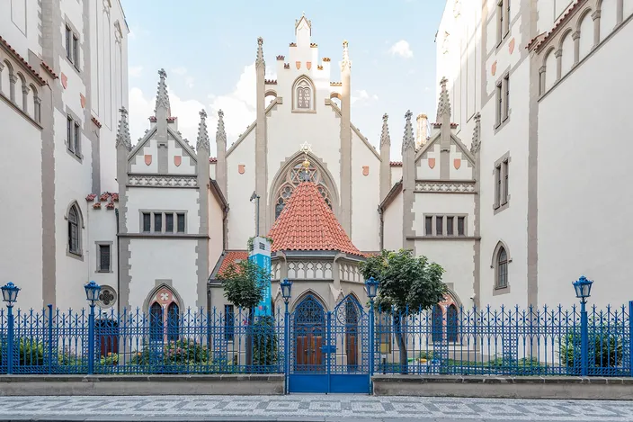 A white neo-Gothic synagogue facade with three stepped gables and slender pinnacles, a small kiosk with a red-tiled pyramid roof standing in front of its central portal, seen behind a blue-painted iron railing with clipped trees on either side in low sunlight.