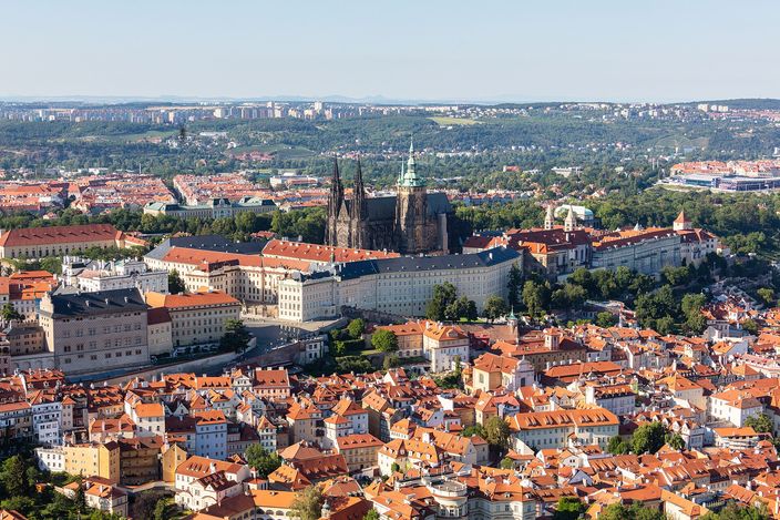 Prague Castle seen from above and to the south: a long pale palace range with a red roof running along a ridge, the dark twin spires and green-capped tower of St Vitus Cathedral rising from its centre, red-tiled roofs stepping down the hillside in the foreground and the flat city running to wooded hills on the horizon.