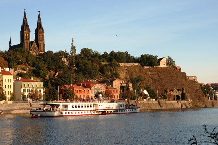 A rocky wooded bluff rising above a river in low evening light, with two dark neo-Gothic spires on the skyline and fortress walls along the crest, pastel houses along the embankment at its foot and a long white excursion steamer moored in front of them.