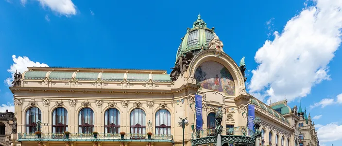A long art nouveau palace of pale stone and cream stucco under a blue sky with white cloud: rows of tall arched windows behind wrought-iron balconies along the front, and at the right-hand end a green copper dome rising above a large semicircular mosaic of figures, with a green and gold ironwork canopy beneath it.