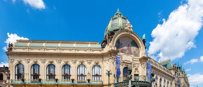 A long art nouveau palace of pale stone and cream stucco under a blue sky with white cloud: rows of tall arched windows behind wrought-iron balconies along the front, and at the right-hand end a green copper dome rising above a large semicircular mosaic of figures, with a green and gold ironwork canopy beneath it.