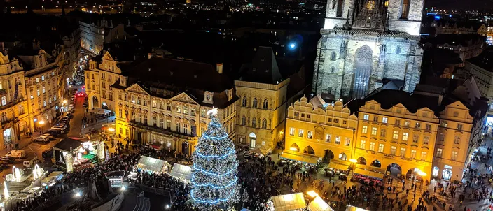 Old Town Square seen from above at night: the lit Christmas tree and the market huts below, the twin spires of the Týn Church rising over the baroque fronts, the city's lights beyond