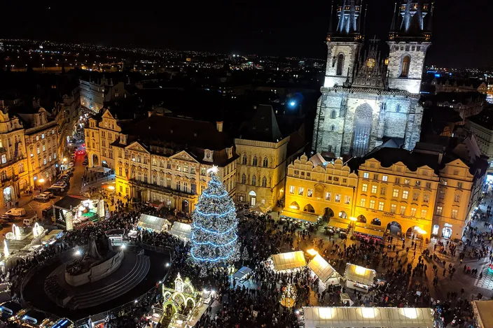 Old Town Square seen from above at night: the lit Christmas tree and the market huts below, the twin spires of the Týn Church rising over the baroque fronts, the city's lights beyond