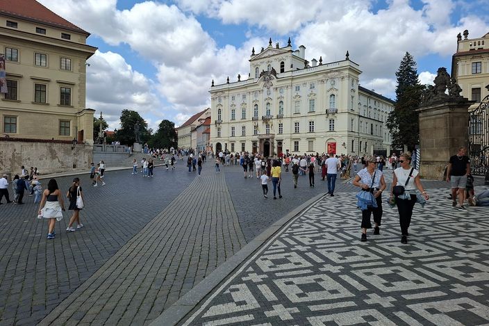 Hradčanské náměstí (Hradčany Square) in Prague, a broad cobbled plaza with a patterned paving mosaic in the foreground, the baroque Archbishop's Palace across the square, and people strolling under a blue sky with clouds.