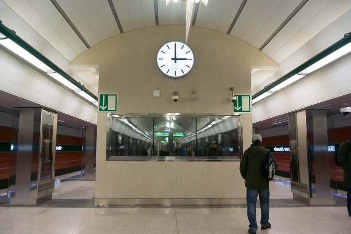 Underground metro platform at Nádraží Veleslavín with a large round white clock on a stone-clad wall, green exit arrows and a man with a backpack waiting