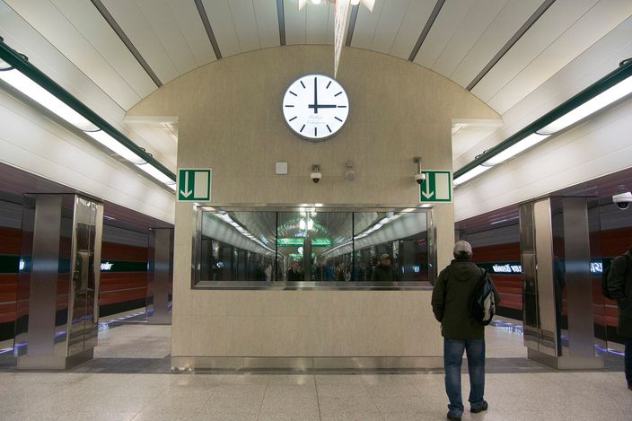 Underground metro platform at Nádraží Veleslavín with a large round white clock on a stone-clad wall, green exit arrows and a man with a backpack waiting