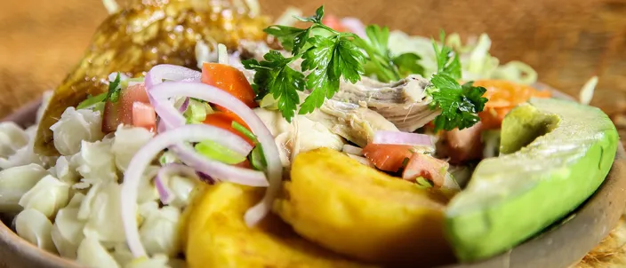 A wooden bowl of hornado: shredded roast pork with a piece of glazed crackling, white mote, fried potato, a curtido of tomato and red onion with coriander, and a wedge of avocado