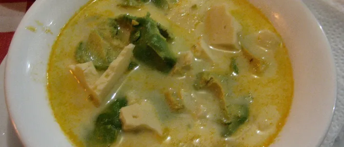 A white bowl of locro de papa: a golden, achiote-tinted potato and milk soup with soft cubes of fresh white cheese and chunks of avocado floating in it