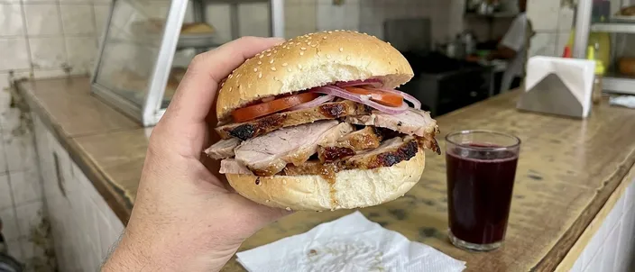 AI-generated image of a sánduche de pernil: a round sesame roll filled with thick slices of roast pork leg with browned edges, tomato and thin rings of red onion, held up in one hand over a shop counter