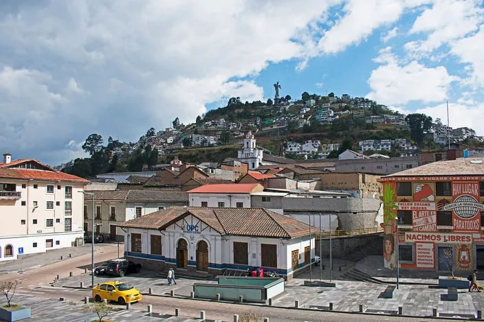 El Panecillo seen from Calle Cuenca: a green hill covered in houses with the tall aluminum winged Virgin on its summit, above the tiled roofs and a small square of the old town