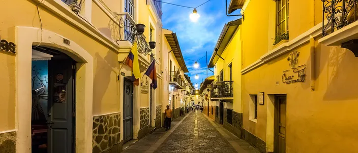 La Ronda at dusk: a narrow cobbled colonial street of yellow and ochre houses with wrought-iron balconies, lanterns and lit doorways