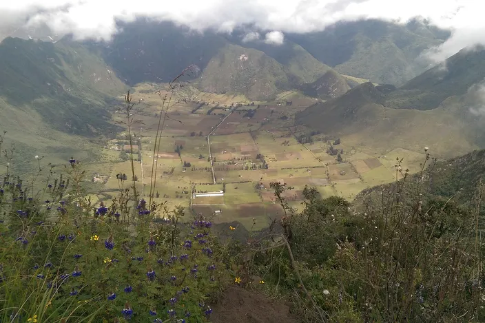 The floor of the Pululahua crater seen from its rim: a patchwork of green and brown fields and a few houses inside a ring of steep, cloud-topped volcanic walls