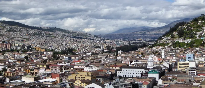 The red-roofed old town of Quito seen from high above, churches and colonial blocks running south along the valley toward green hills under a cloudy sky