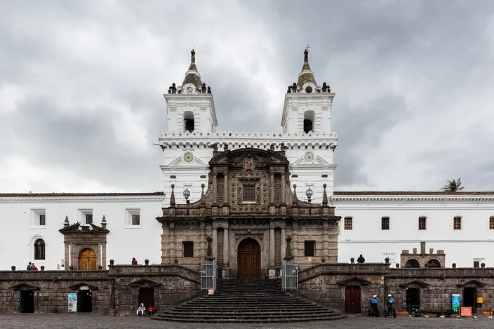 The white twin-towered facade of the church and convent of San Francisco above its wide stone steps and the paved plaza in front, under a grey sky