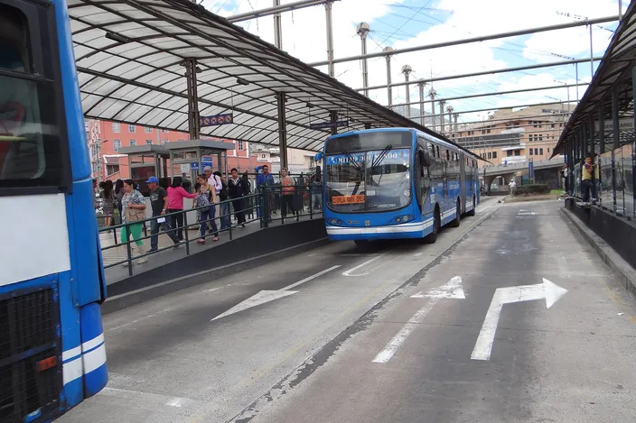 A blue articulated bus pulling into the covered platforms of a busy bus rapid transit station in Quito, passengers waiting along the barrier
