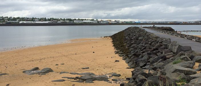 A curve of imported golden sand meeting a long arm of dark volcanic boulders that walls the cove off from the open bay at Nautholsvik, with the low buildings of Reykjavik along the far shore under a heavy grey sky.