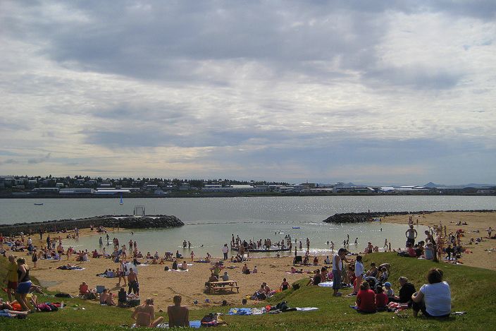The lagoon at Nautholsvik full of bathers, enclosed by two arms of dark rock, with families spread across the golden sand and the grass bank above, and the city and its low mountains on the horizon.