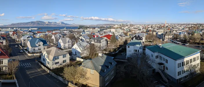 Elevated panorama over central Reykjavík: rows of small houses with red, green and blue metal roofs, the bay and snow-flecked mountains in the distance under a bright partly cloudy sky.