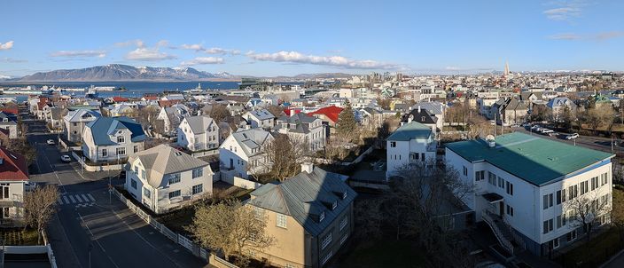 Elevated panorama over central Reykjavík: rows of small houses with red, green and blue metal roofs, the bay and snow-flecked mountains in the distance under a bright partly cloudy sky.