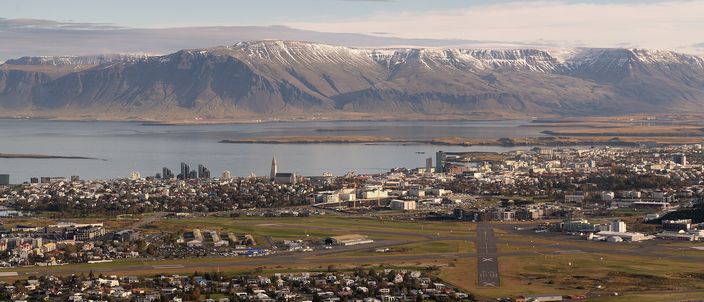 Reykjavík seen from the air in low afternoon light: the low-rise city spread along the shore with the spire of Hallgrímskirkja rising from the middle of it, the domestic airport runways in the foreground, the bay beyond, and the long snow-streaked flat-topped ridge of Esja filling the far side.