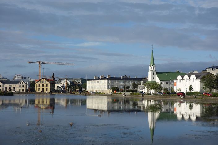 Tjörnin, the lake in central Reykjavík, mirror-flat under a cloudy sky, with the green-spired Fríkirkjan church, a row of pale houses and a construction crane reflected in the water and ducks on the surface.
