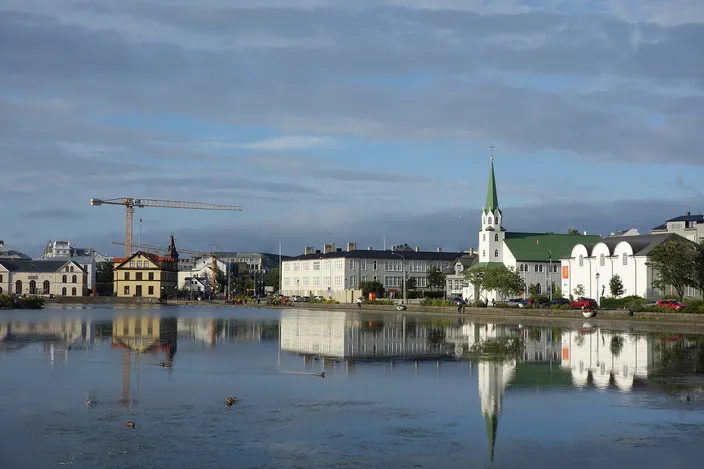 Tjörnin, the lake in central Reykjavík, mirror-flat under a cloudy sky, with the green-spired Fríkirkjan church, a row of pale houses and a construction crane reflected in the water and ducks on the surface.