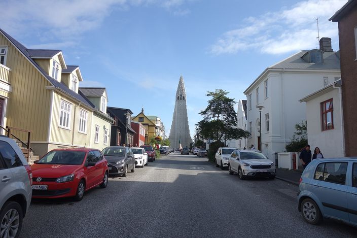 Looking up Skólavörðustígur in Reykjavík between rows of timber and metal-clad houses in yellow, red and white, cars parked along both kerbs, with the stepped grey concrete tower of Hallgrímskirkja closing the end of the street.