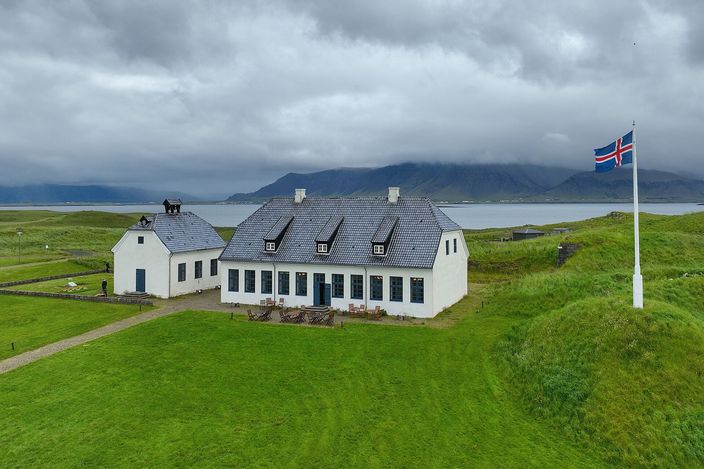 Aerial view of a long low white house and a small white church with dark slate roofs standing on the green island of Viðey, an Icelandic flag flying from a mast to the right, the sound and the cloud-covered Esja range behind them.