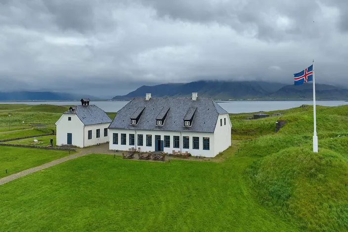 Aerial view of a long low white house and a small white church with dark slate roofs standing on the green island of Viðey, an Icelandic flag flying from a mast to the right, the sound and the cloud-covered Esja range behind them.