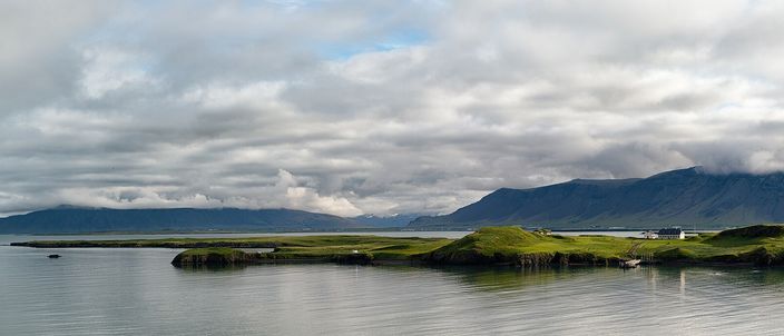 A wide view across calm grey-blue water to the low green island of Viðey, a few small white buildings strung along its ridge, with the long cloud-capped Esja range filling the horizon behind it.