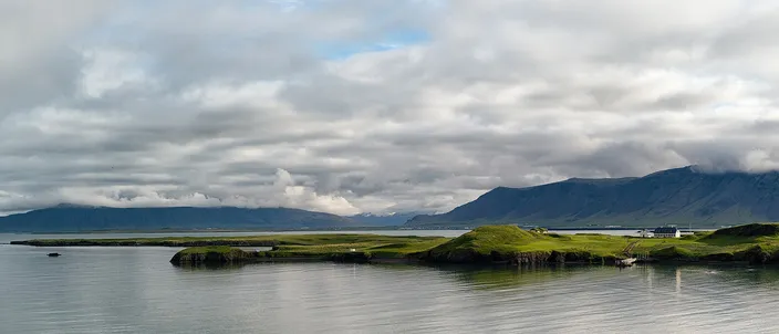 A wide view across calm gray-blue water to the low green island of Viðey, a few small white buildings strung along its ridge, with the long cloud-capped Esja range filling the horizon behind it.