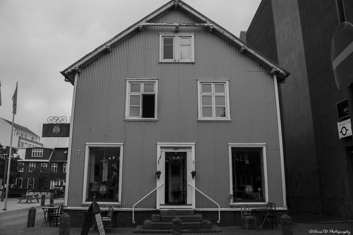 Black-and-white photograph of a two-storey corrugated-iron-clad Reykjavík townhouse with a steep gable, tall windows, a central white door reached by steps, and cafe tables and chairs set out on the pavement in front