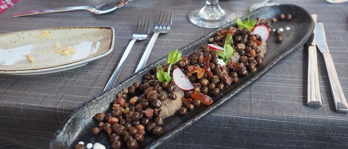A long dark plate of Latvian grey peas with bacon: heaped grey-brown field peas mixed with diced fried pork, garnished with radish and pea shoots, on a restaurant table.
