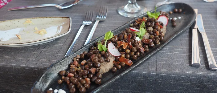 A long dark plate of Latvian gray peas with bacon: heaped gray-brown field peas mixed with diced fried pork, garnished with radish and pea shoots, on a restaurant table.