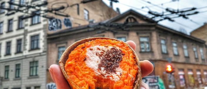 A hand holding a sklandrausis with its bright orange carrot top, in a Riga street with tram wires overhead