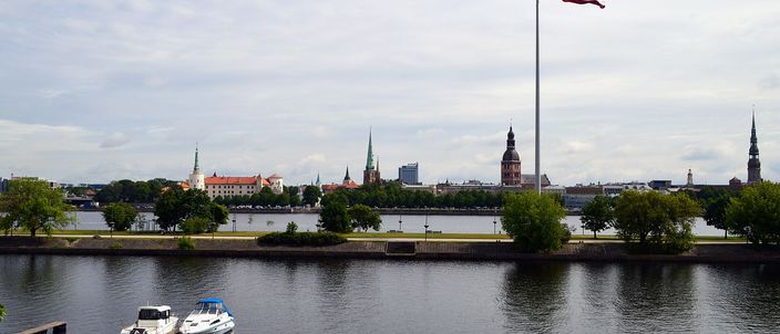 The Riga skyline across the wide Daugava river, with the spires of St Peter's, the Dome Cathedral and the castle rising behind a large Latvian flag on a mast, two small boats moored at a pontoon in the foreground.