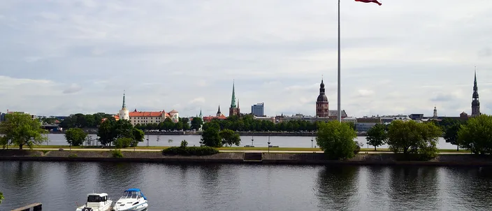 The Riga skyline across the wide Daugava river, with the spires of St Peter's, the Dome Cathedral and the castle rising behind a large Latvian flag on a mast, two small boats moored at a pontoon in the foreground.