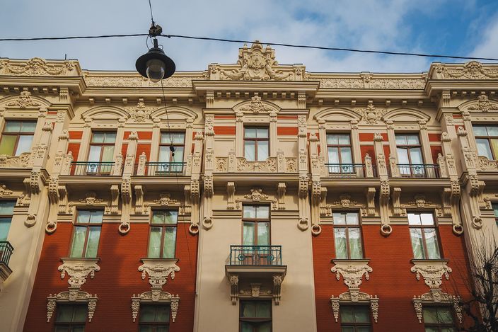 The upper storeys of an Art Nouveau apartment block in Riga photographed from the street below, its red and cream facade crowded with sculpted masks, scrolls and floral plasterwork around tall windows and a small iron balcony, a street lamp hanging on a wire against a blue sky.
