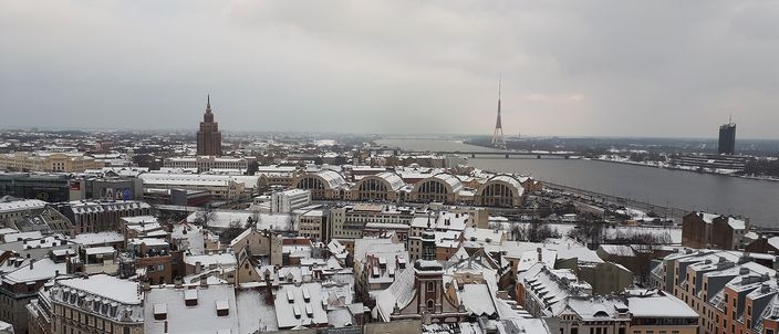 Snow-covered red-tiled rooftops of Riga's old town seen from a church tower, with the long arched roofs of the Central Market pavilions and the wide Daugava river beyond, and the spired Academy of Sciences tower and a slender TV tower on the skyline under a grey winter sky.