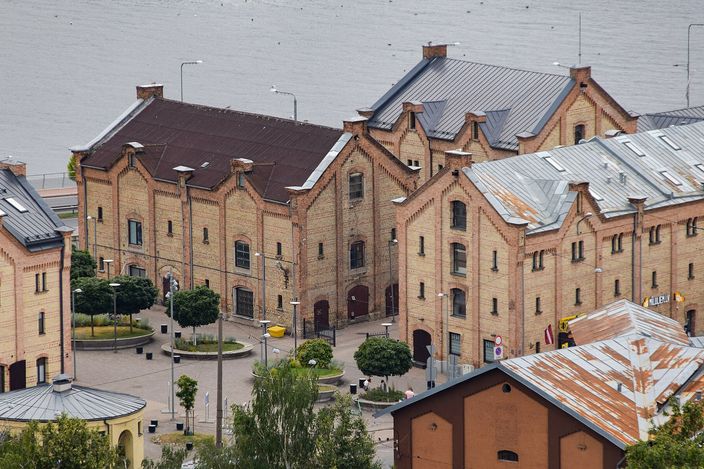 A block of nineteenth-century warehouses in Riga seen from above: long two- and three-storey buildings of pale yellow brick with stepped gable ends and metal roofs, arranged around a paved square planted with clipped trees and low hedges, the grey water of the Daugava filling the top of the frame.