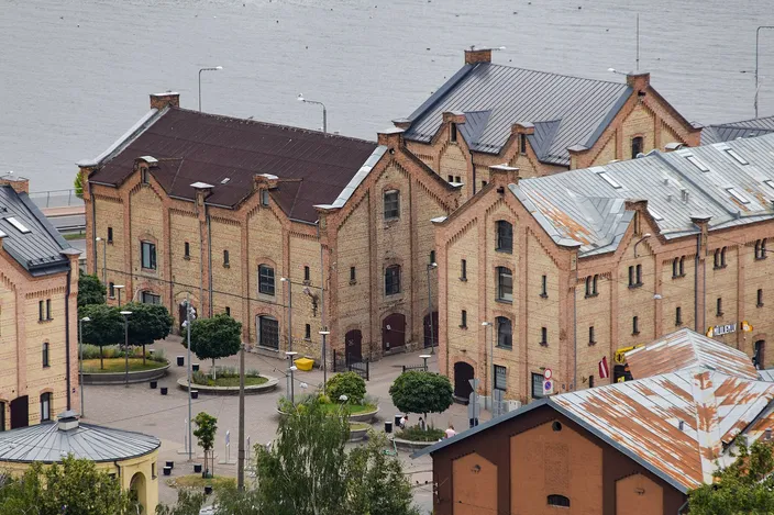 A block of nineteenth-century warehouses in Riga seen from above: long two- and three-story buildings of pale yellow brick with stepped gable ends and metal roofs, arranged around a paved square planted with clipped trees and low hedges, the gray water of the Daugava filling the top of the frame.