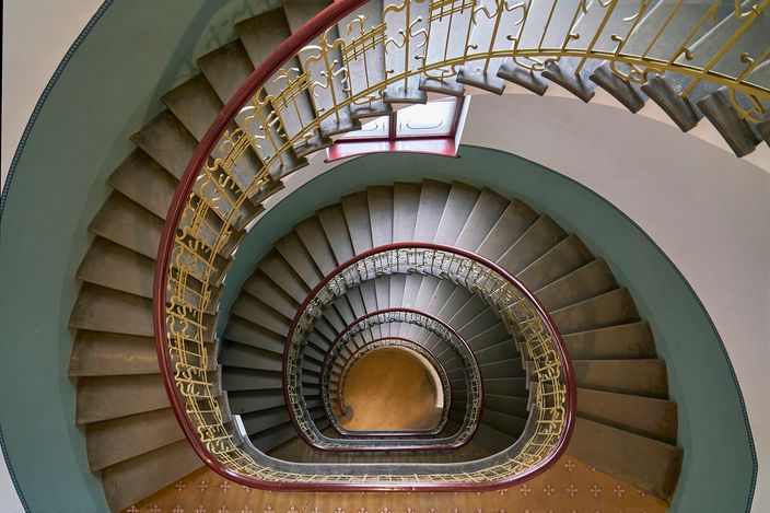 An Art Nouveau spiral staircase photographed from the top landing looking straight down, its stone treads winding round three full turns to a small landing far below, with a dark red timber handrail and an ornate gilded wrought-iron balustrade of curling stems against pale green walls.