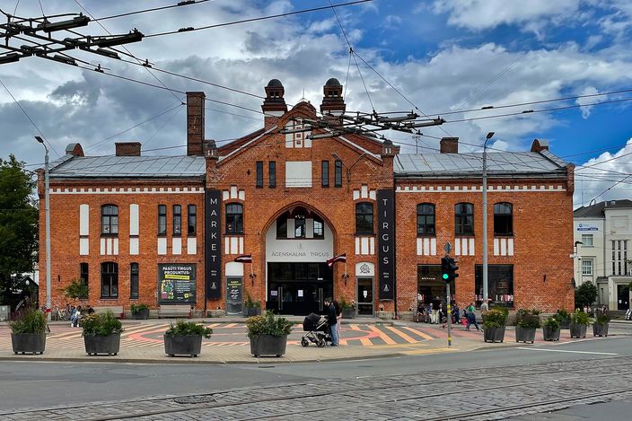 A red brick market hall in Riga seen across a tram-lined street: a symmetrical two-storey building with a central pointed arch, a stepped gable and two brick turrets, MARKET and TIRGUS lettered vertically on black panels either side of the entrance, planted tubs and a striped pedestrian crossing in front, tram wires overhead against a blue sky with heavy white cloud.