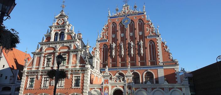 The House of the Blackheads on Riga's town hall square in low sunlight: two stepped gables side by side, the right one tall red brick with white stone statues in niches, a blue and gold clock face and a gilded weathervane on its apex, the left one narrower and paler, a group of people standing on the paving in front of the entrance under a clear blue sky.