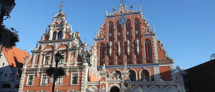 The House of the Blackheads on Riga's town hall square in low sunlight: two stepped gables side by side, the right one tall red brick with white stone statues in niches, a blue and gold clock face and a gilded weathervane on its apex, the left one narrower and paler, a group of people standing on the paving in front of the entrance under a clear blue sky.