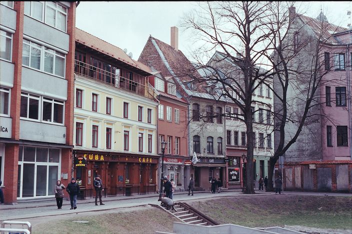 A cobbled Old Town square in Riga lined with pastel gabled townhouses and a café, bare trees and people walking under an overcast late-winter sky