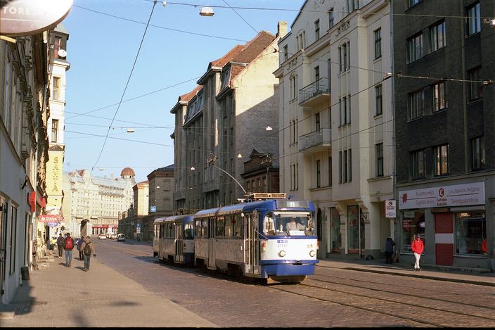A blue-and-white Tatra T3 tram runs down Matīsa iela in Riga between five-storey apartment buildings, overhead wires above, pedestrians on the pavement and a Latvian flag on a building at right.