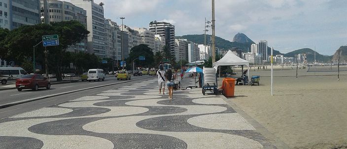 The black-and-white wave-patterned mosaic promenade of Copacabana running beside a wall of apartment blocks and hotels, with a volleyball net, a vendor's tent and a dog-walker on the pavement, and the open Atlantic and a distant granite peak beyond the sand
