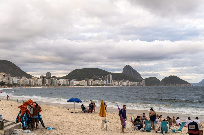 A wide stretch of Copacabana sand seen from Posto 5 on an overcast afternoon — beach umbrellas, deckchairs and a vendor's tent in the foreground, whitewater breaking along the shoreline, and a twin-peaked mountain beyond the water